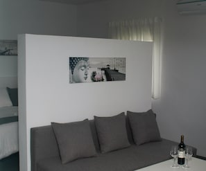 Een kamer met een grijze bank, een aan de muur hangend schilderij, een fles wijn en een raam met gordijnen.