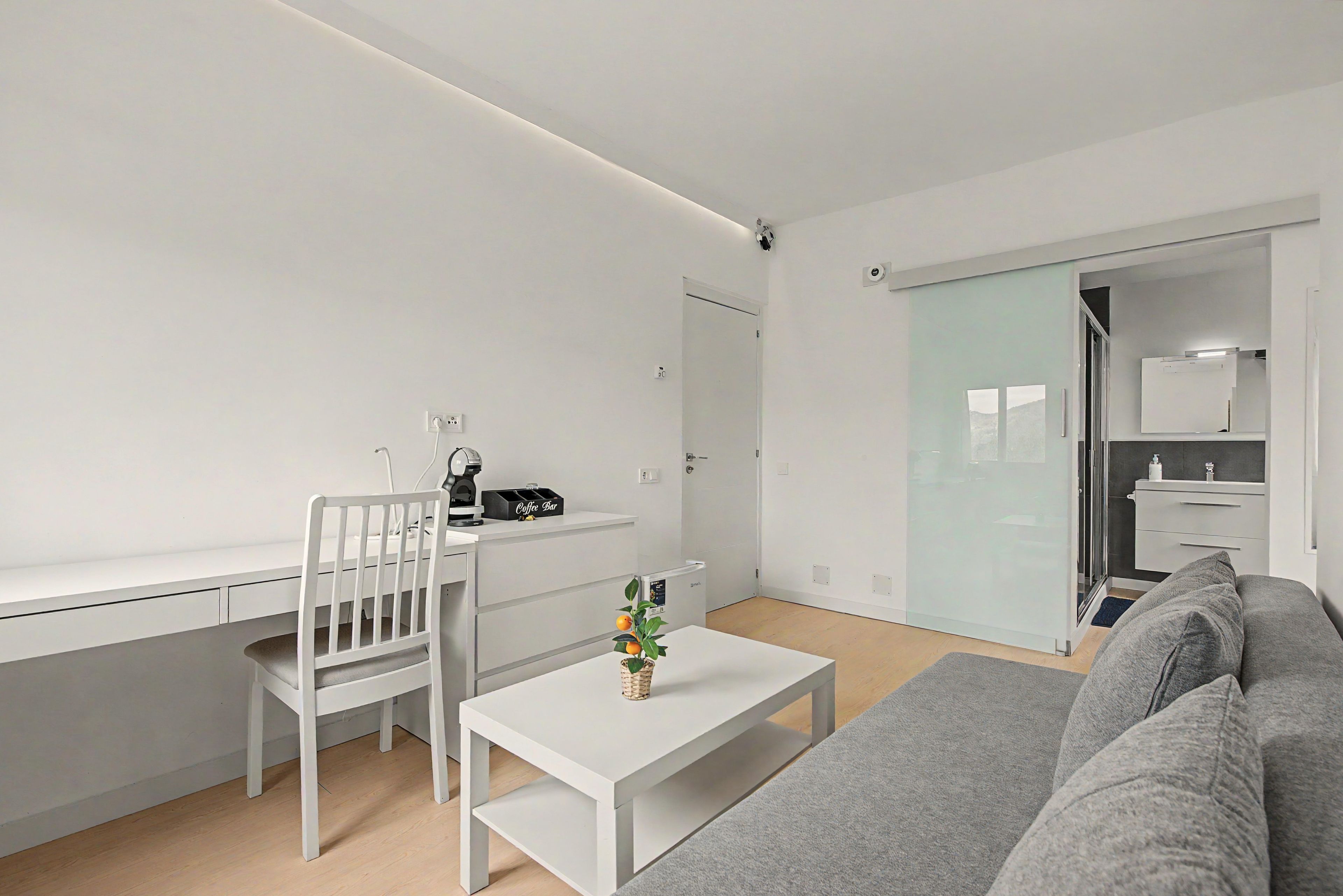 Ett minimalistiskt vardagsrum med ett vitt skrivbord, en grå soffa och ett litet bord med en växt.
