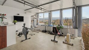 A gym with exercise equipment, a television, and a view of a hilly landscape through large windows. - Jingtu Wellness & Spa Vairochana (Romo Cuevas)
