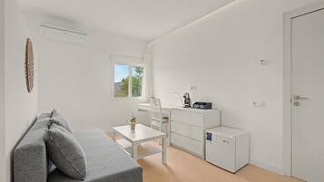 A modern, minimalist room with a grey sofa, a small white table, a coffee machine, and a compact refrigerator.