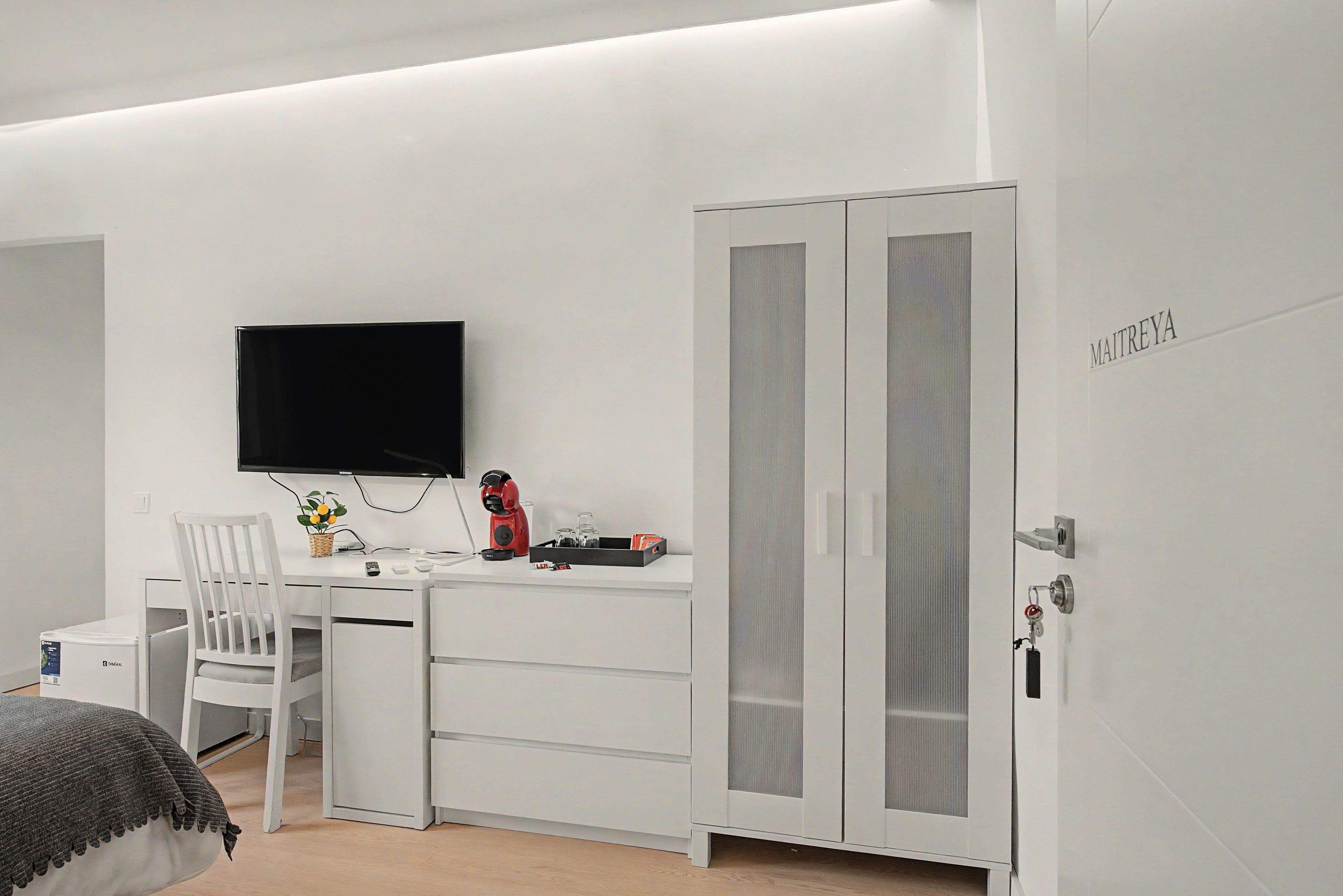 Un dormitorio minimalista con una cama, un escritorio con televisor, un armario y una mesita de noche con lámpara y planta.
