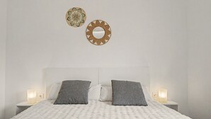 A bedroom with a white quilted bedspread, two pillows, two wall-mounted lamps, and two decorative wall hangings. - Jingtu Wellness & Spa Maitreya (Romo Cuevas)