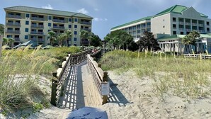 Ein Strandhotel mit einer Holzpromenade, die zum Strand führt, umgeben von Sand und Vegetation.