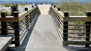 Ein hölzerner Steg führt zu einem Strand mit Dünen und Strandsonnenschirmen im Hintergrund.