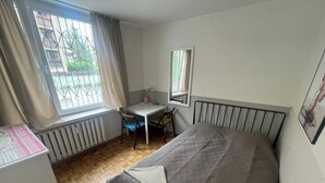 A bedroom with a bed, a desk, a chair, a radiator, and a window with curtains. - Lama Rooms (Warsaw)