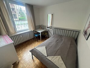 A bedroom with a bed, a desk, a chair, a radiator, and a window with curtains. - Lama Rooms (Warsaw)