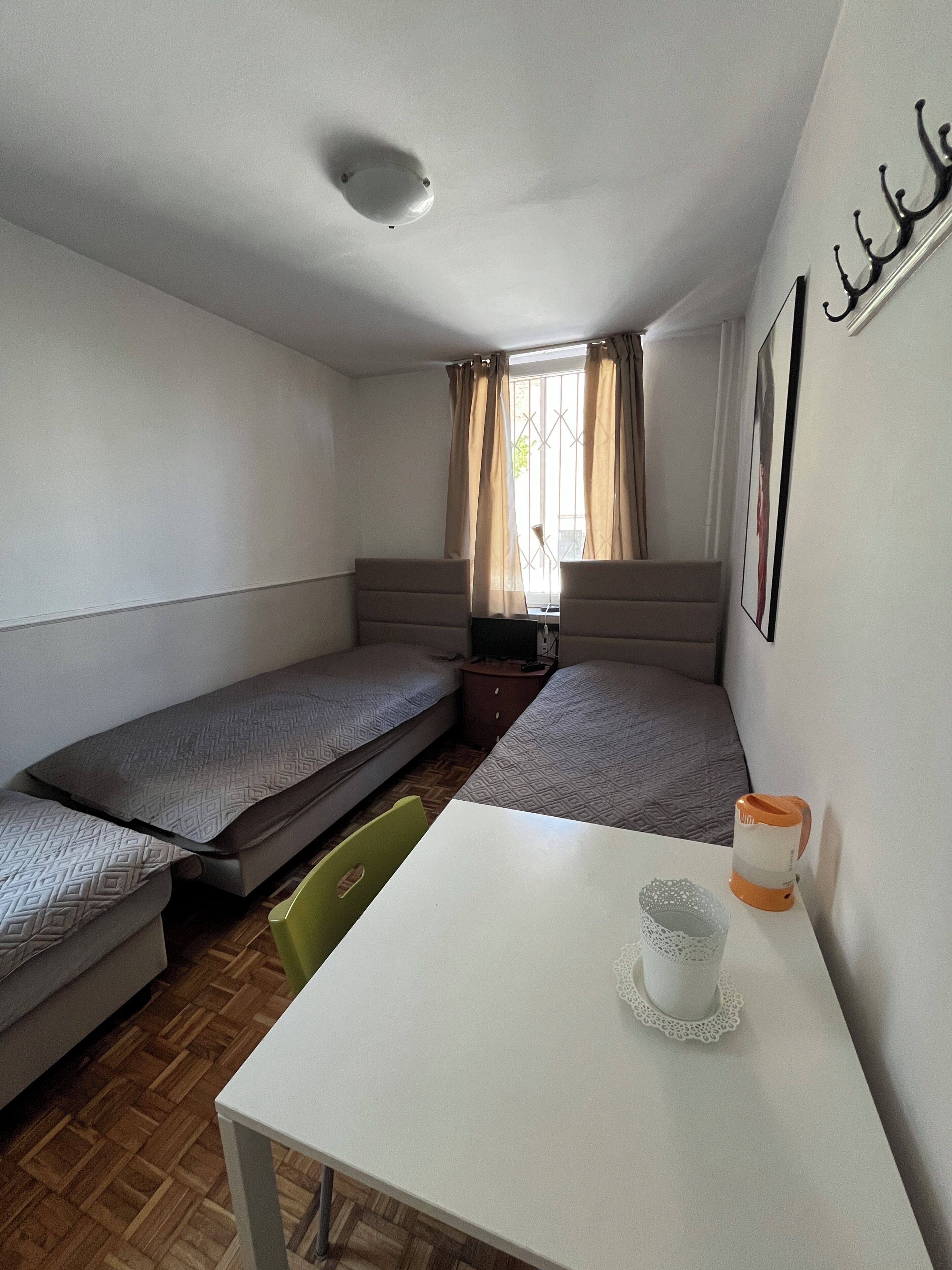 Una habitación pequeña y amoblada con dos camas individuales, una mesa y un ventanal con cortinas.