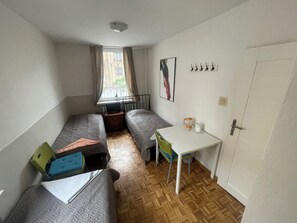 Una habitación pequeña con dos camas individuales, una mesa, una silla, una ventana con cortinas, un radiador, un cuadro en la pared y una puerta.