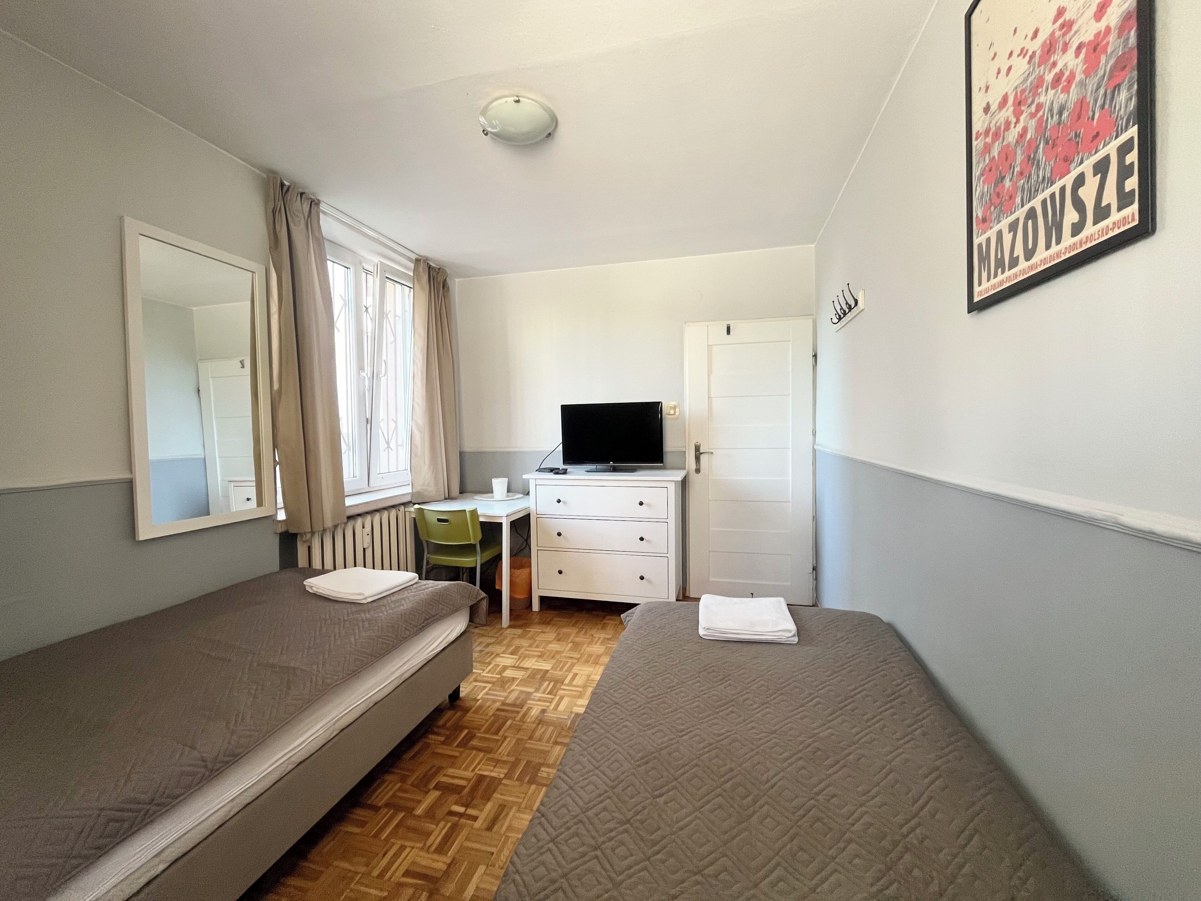 A small, single-room accommodation with two beds, a desk, a chair, a television, and a radiator.