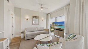 A modern living room with a sofa, a coffee table, and a TV on a stand, with a view of a beach and buildings. - Marina Majesty 7110/12-Lockout Combo-Sleeps 6 (Miramar Beach)