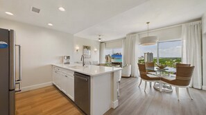 A modern kitchen with a central island, a dining area with a glass table, and a view of a beach and cityscape. - Marina Majesty 7110/12-Lockout Combo-Sleeps 6 (Miramar Beach)