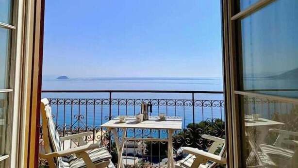 A balcony with a table and chairs, a view of the sea, and a metal railing. - Hostdomus - Bay Apartment (Laigueglia)