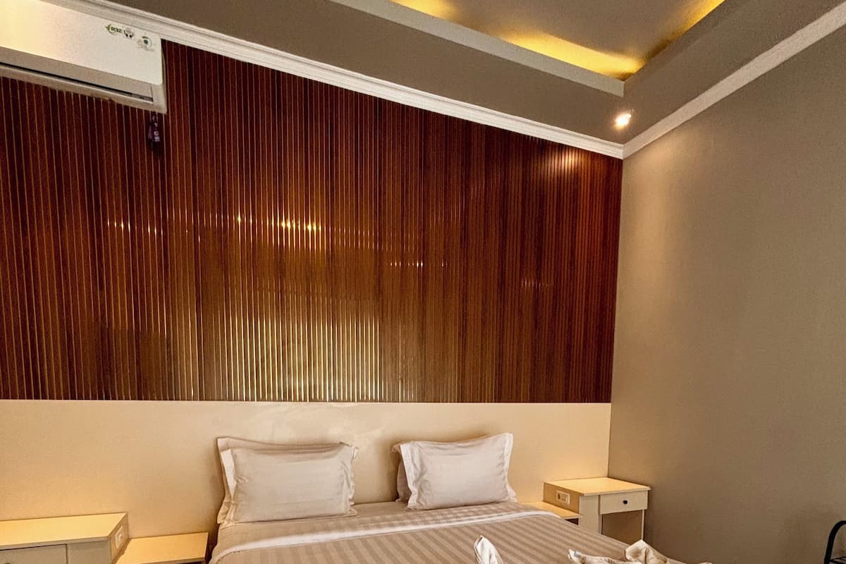 A modern hotel room with a large bed, two bedside tables, a wooden panel wall, and an air conditioning unit.