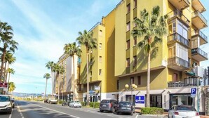 Un immeuble jaune à plusieurs étages, doté de balcons, avec des voitures garées et des palmiers.