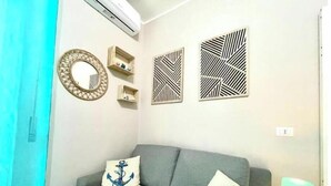 Un canapé gris avec deux coussins, l’un orné d’un motif d’ancre et l’autre d’un motif d’hippocampe. On y trouve également des panneaux décoratifs fixés au mur et un miroir.