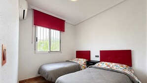 Una stanza con due letti, una finestra con tende rosse, un termostato e un punto luce a parete.
