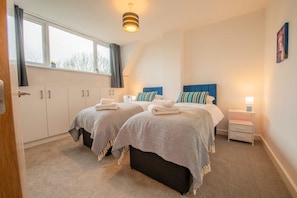 A bedroom with two beds, a window, a nightstand, and a door. - Stylish Home Near Scotch Corner For Work Leisure (Richmond)