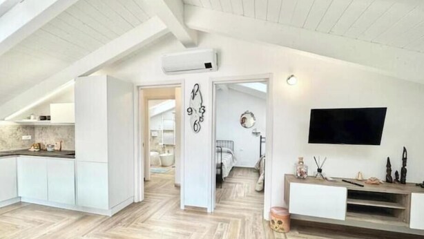 A modern loft with a high ceiling, exposed beams, a kitchen with white cabinetry, a living area with a TV, and a bedroom with a bed and a bathroom in the background. - Hostdomus - Bellavista Suite (Pietra Ligure)