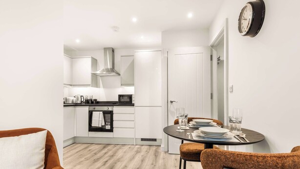 Een moderne keuken met witte kasten, een zwart aanrecht en een roestvrijstalen afzuigkap. Er is een eethoek met een ronde tafel, twee stoelen en een klok aan de muur.