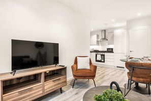 A modern living room with a flat-screen TV, wooden entertainment unit, brown armchair, dining table with chairs, and a kitchen in the background. - Bright & Contemporary 1 Bed Apartment in Leeds (Leeds)