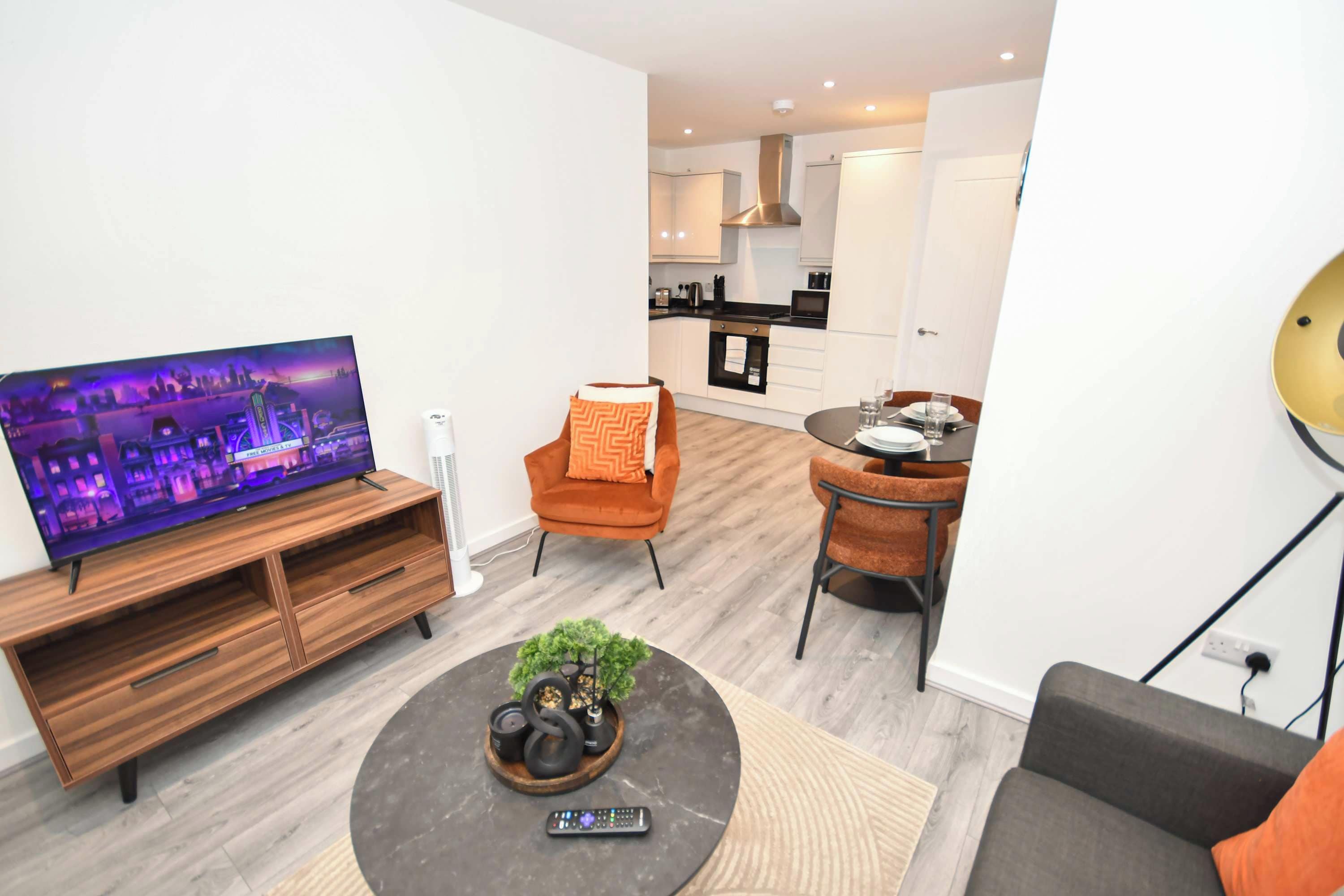 Een moderne woonkamer met een flatscreen televisie, een bijzettafeltje met decoratieve objecten, een bank en een eethoek met een tafel en stoelen.