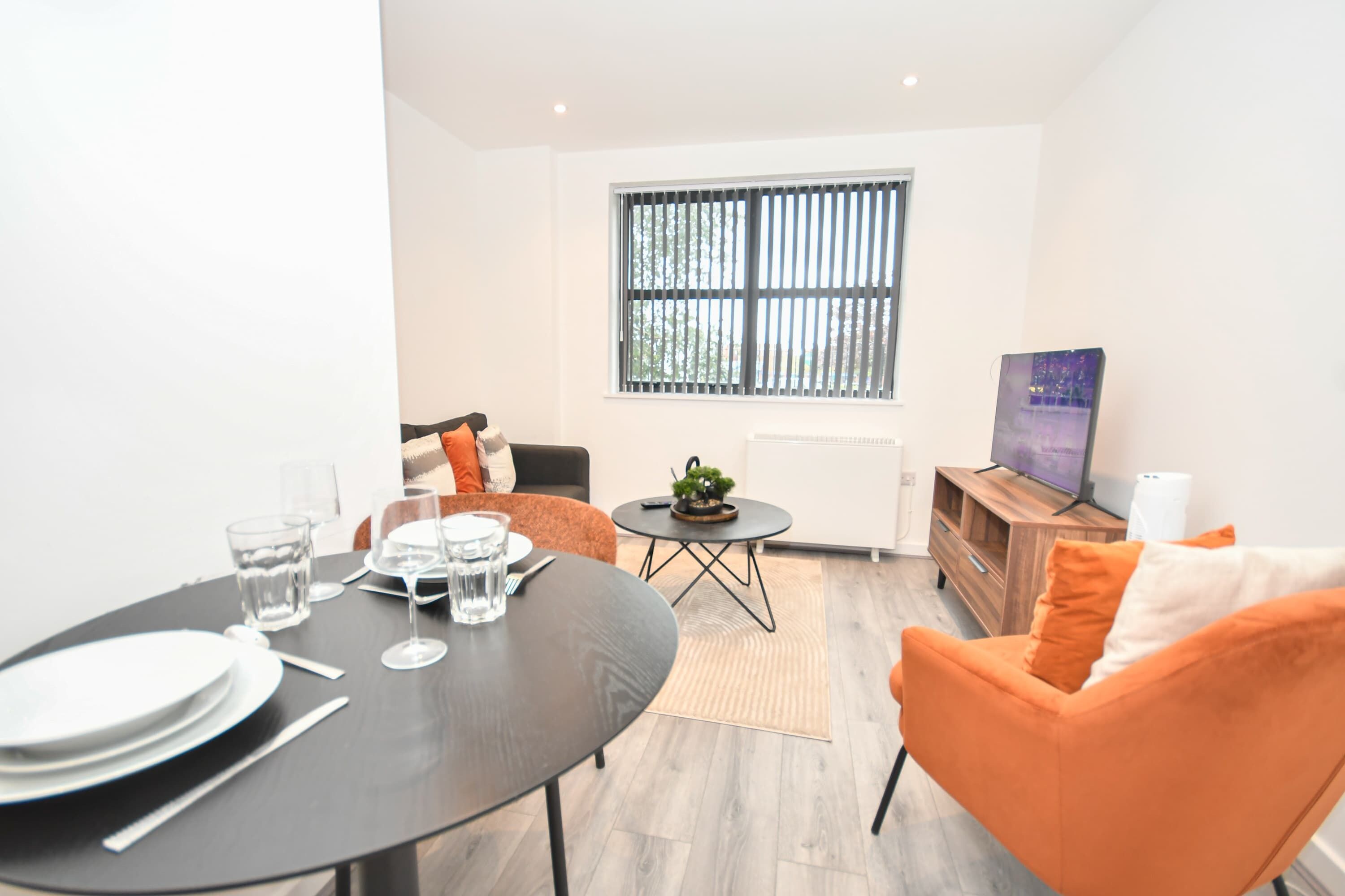 A modern living room with a dining table set for two, a sofa, a TV, and a window with blinds.