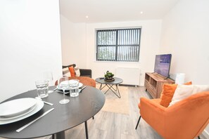 A modern living room with a dining table set for two, a sofa, a TV, and a window with blinds. - Bright & Contemporary 1 Bed Apartment in Leeds (Leeds)