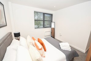 A hotel room with a bed, a bedside table with a lamp, a window with blinds, a mirror, and a radiator. - Bright & Contemporary 1 Bed Apartment in Leeds (Leeds)