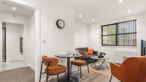 Een moderne woonkamer met een ronde eettafel, twee oranje stoelen, een grijze bank en een groot raam met verticale jaloezieën.
