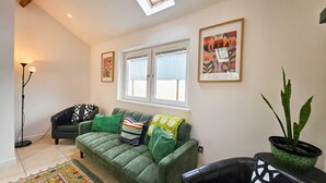 A living room with a green sofa, a floor lamp, a window with curtains, and a potted plant. - Buttercup - 2 Bed Cottage - Red Forest Retreats (Neath)