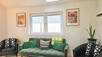 A living room with a green sofa, two armchairs, a window with blinds, and two framed artworks.