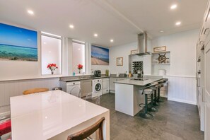 A modern kitchen with a central island, bar stools, a washing machine, and a large window with a beach view. - Finest Retreats | Wallingbrook Lodge (Chulmleigh)
