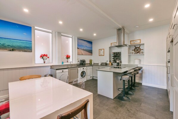 A modern kitchen with a central island, bar stools, a washing machine, and a large window with a beach view. - Finest Retreats | Wallingbrook Lodge (Chulmleigh)