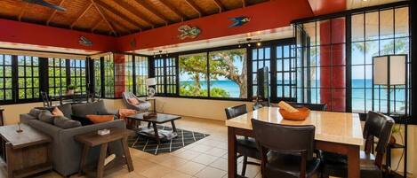 A spacious room with a wooden ceiling, large windows offering a view of the ocean, a dining area with a table and chairs, and a seating area with a sofa and armchairs.