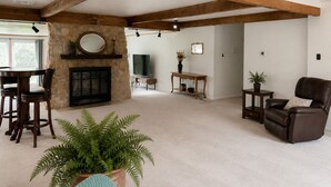 Ein großzügiges Wohnzimmer mit Kamin aus Stein, Holzbalken, einem Essbereich mit Stühlen, einer Couch und einem Topf-Farn.
