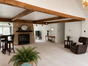 Ein großzügiges Wohnzimmer mit Kamin aus Stein, Holzbalken, einem Essbereich mit Stühlen, einer Couch und einem Topf-Farn.