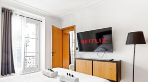 Ein Hotelzimmer mit Bett, Wandfernseher, Nachttisch, einem Fenster mit Vorhängen und Blick nach draußen.