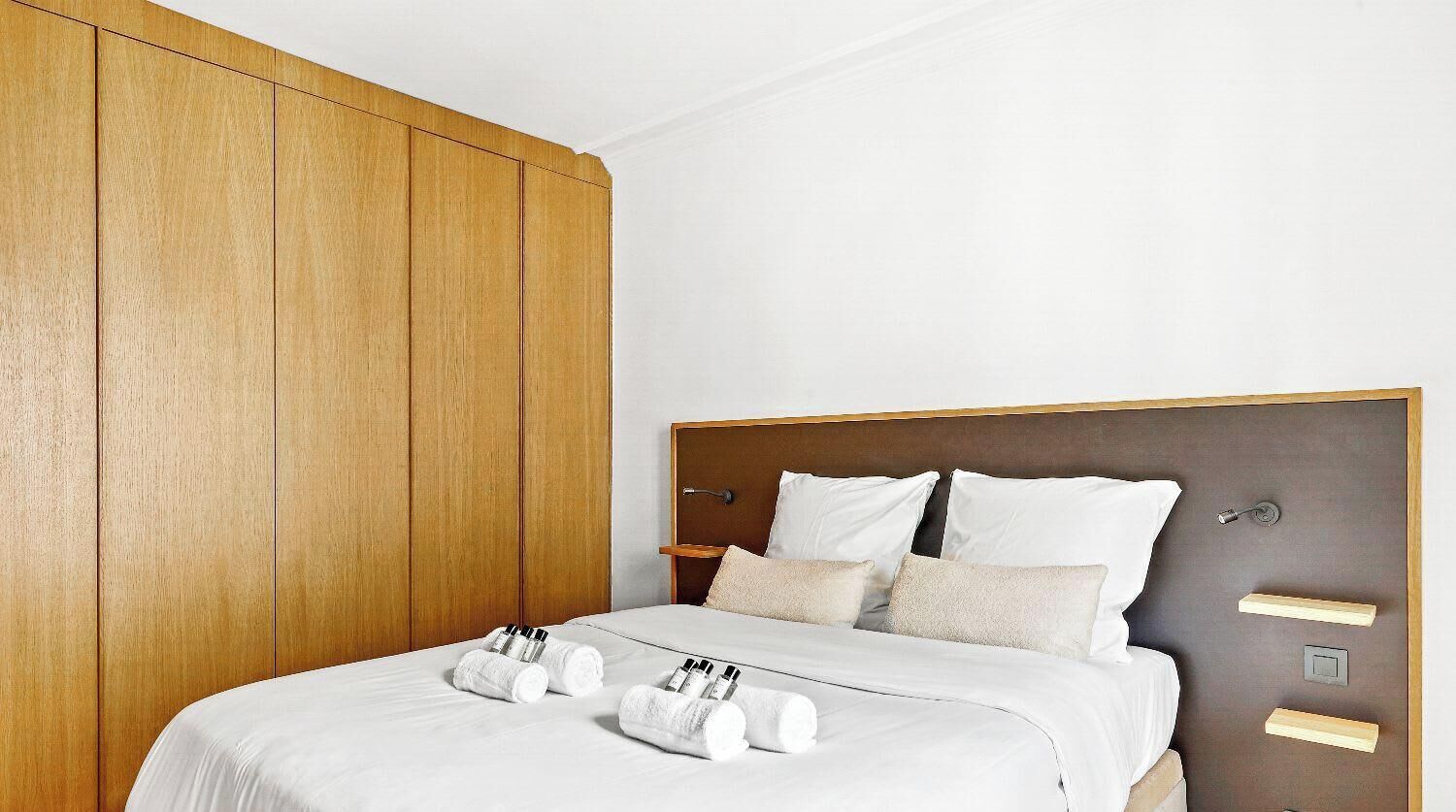 Een hotelkamer met een houten garderobe, een bed met witte lakens en een nachtkastje met toiletartikelen.
