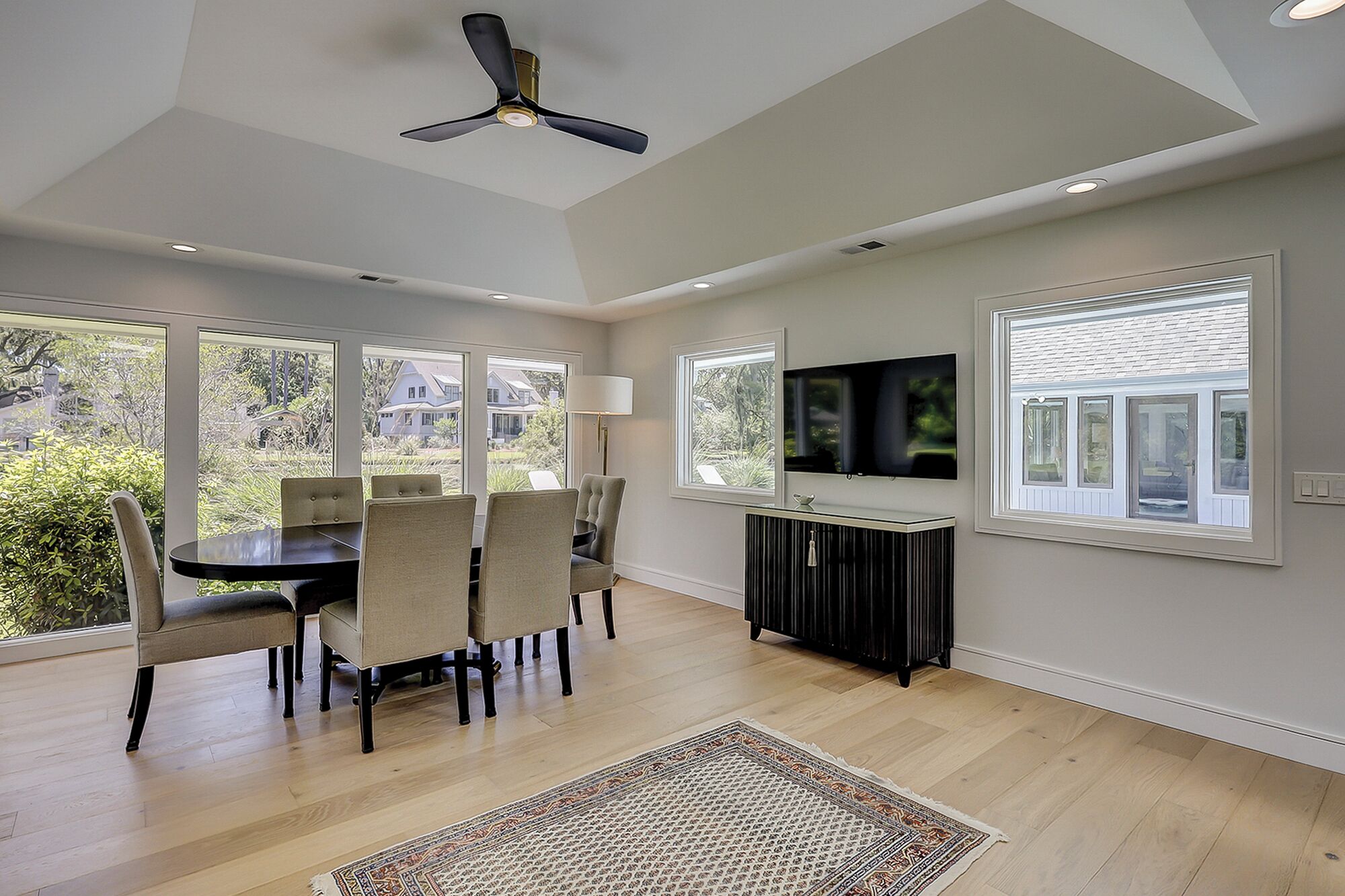 A dining area with a table and chairs, a ceiling fan, a TV on the wall, and large windows.