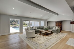 Ein großzügiges Wohnzimmer mit einer breiten Eckcouch, einem Couchtisch und einem weißen Sessel. Ein großes Fenster bietet Blick auf einen Swimmingpool und Gartenmöbel.