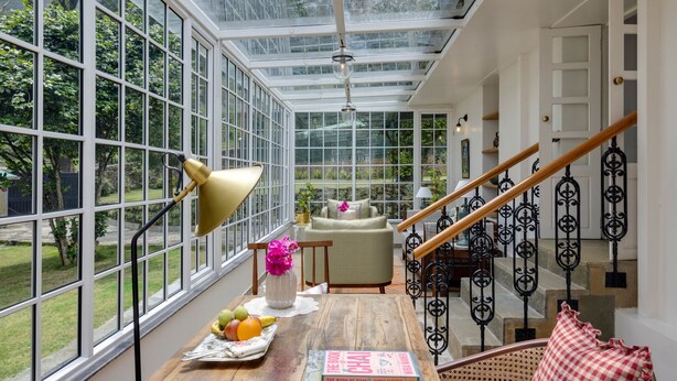 A conservatory with large windows, a wooden table with books and fruit, a chair, and a staircase with wrought iron railings. - Singell Tea Estate- CGH Earth Experience (Kurseong)