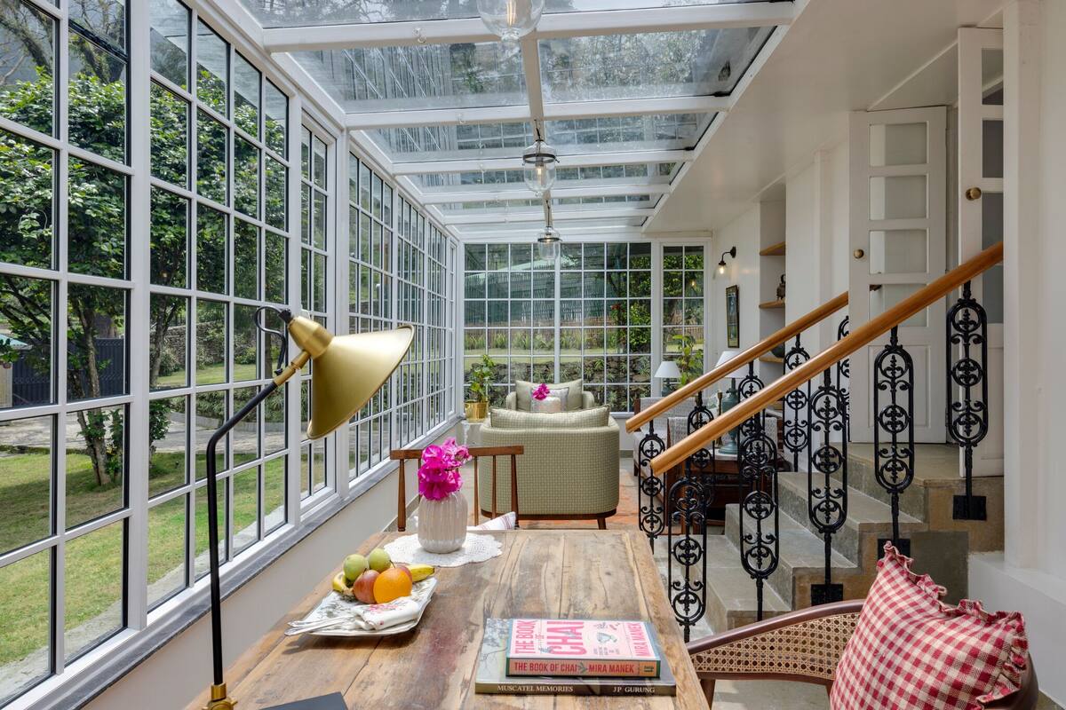 A conservatory with large windows, a wooden table with books and fruit, a chair, and a staircase with wrought iron railings.