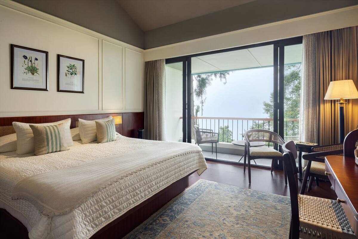 A hotel room with a large bed, a balcony with chairs, and framed botanical prints on the wall.