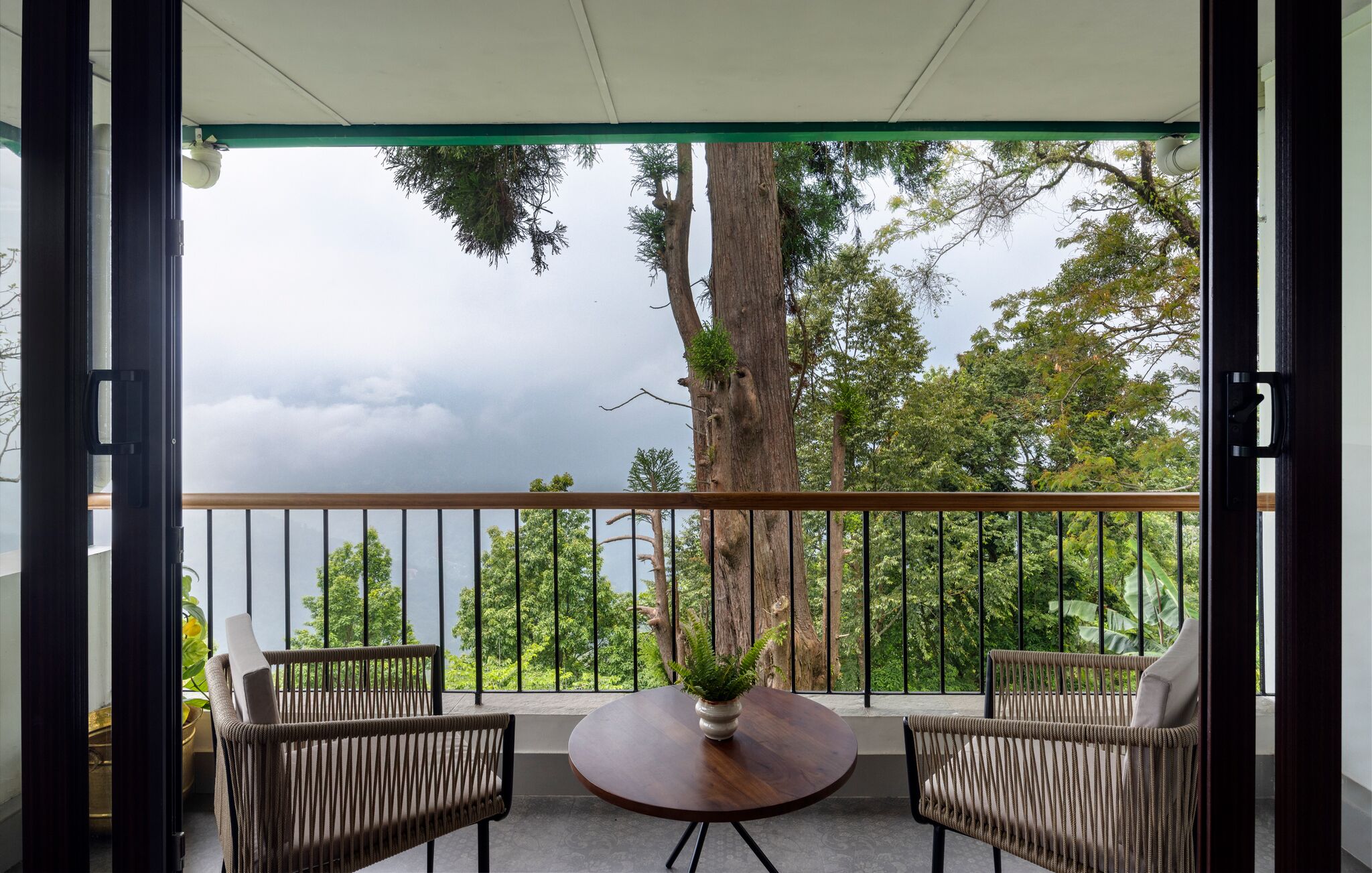 A balcony with two wicker chairs, a round table with a plant, and a view of trees and clouds.