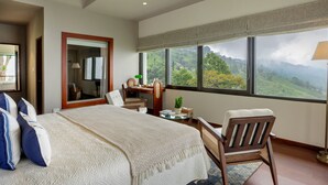A bedroom with a large bed, a desk, a chair, and a view of a mountainous landscape through large windows. - Singell Tea Estate- CGH Earth Experience (Kurseong)
