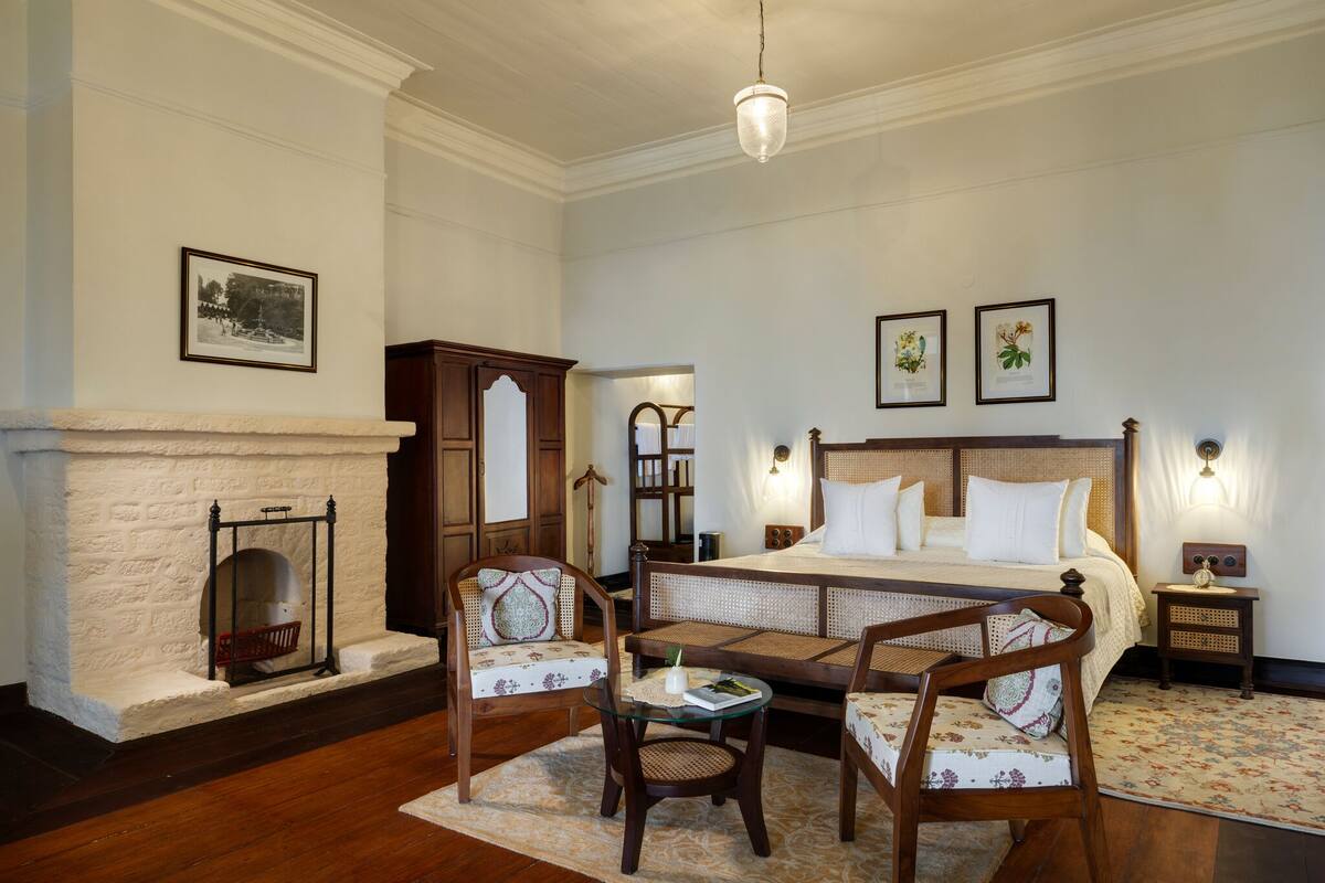 A bedroom with a large bed, two chairs, a small table, a fireplace, and framed pictures on the wall.
