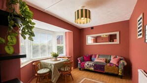 Una habitación acogedora con una mesa de comedor, sillas, un sofá colorido, una planta colgante y una lámpara en el techo.