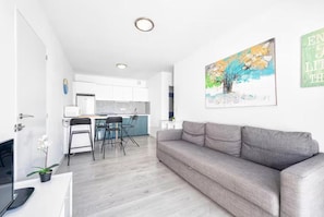 A modern living room with a grey sofa, a dining area with chairs, a kitchen in the background, and a painting on the wall.