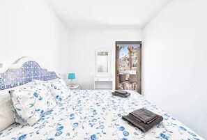 Une chambre à coucher avec un lit, une table de chevet, un miroir et une vue sur un bâtiment par une porte ouverte.