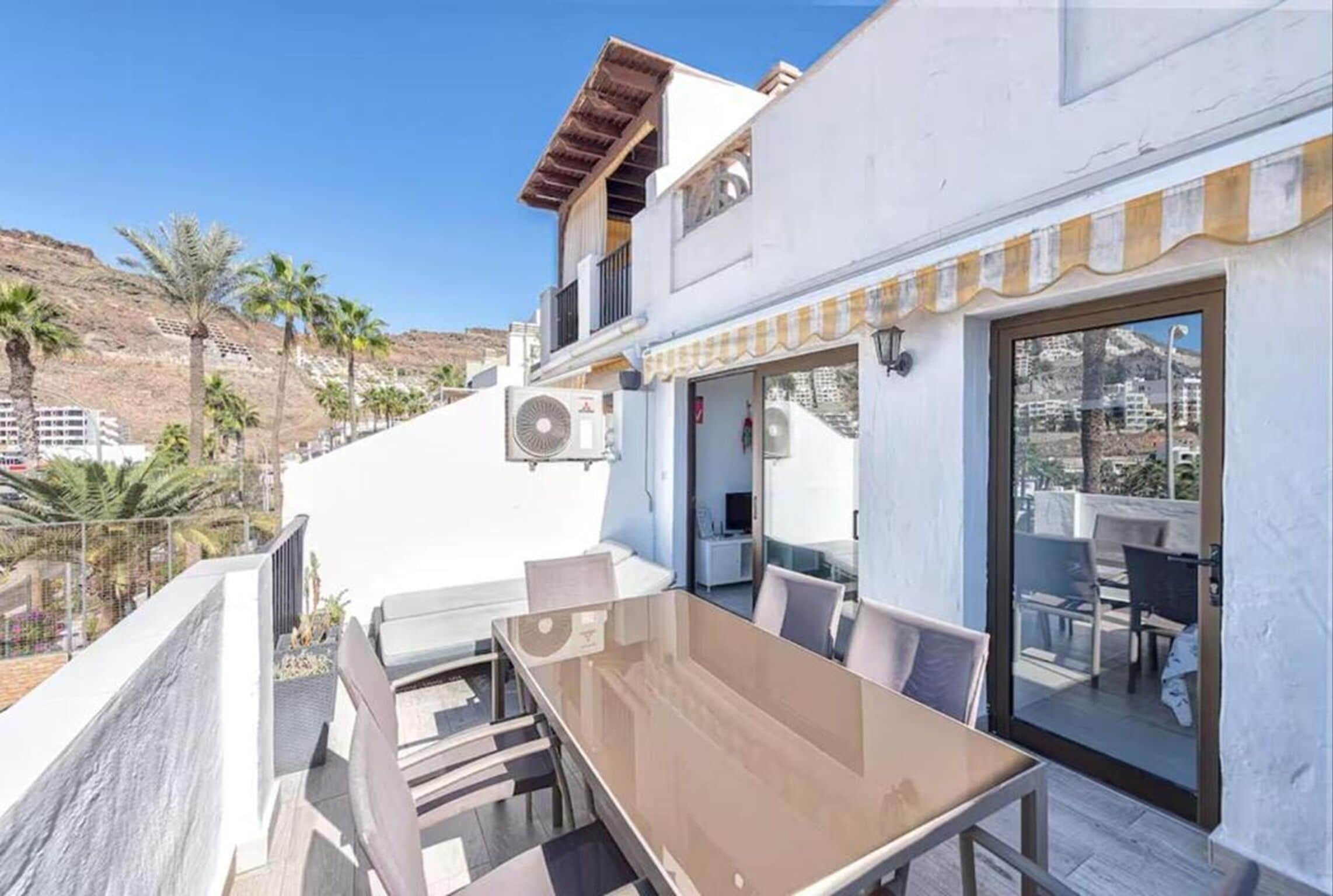 A modern outdoor patio with a glass table and chairs, a white building with a balcony, and a clear view of palm trees and hills in the background.